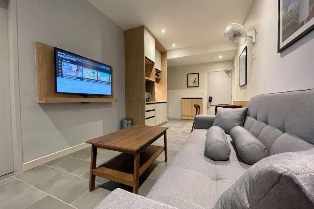 Een moderne woonkamer met een flatscreen televisie, een grijze bank, een houten salontafel en een keuken in de achtergrond.