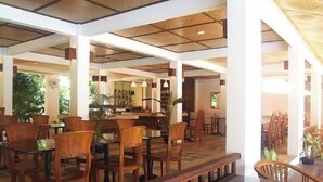 Um espaço amplo e interno com mesas e cadeiras de madeira, um bar central e grandes janelas que permitem a entrada de luz natural.