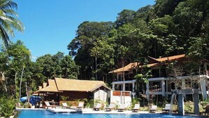 Área de piscina com espreguiçadeiras, um pequeno edifício e uma cascata.