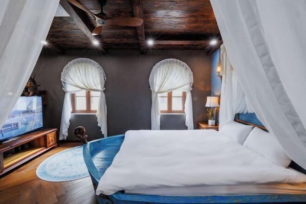 A bedroom with a bed, two windows with curtains, a television, a nightstand, and a lamp.