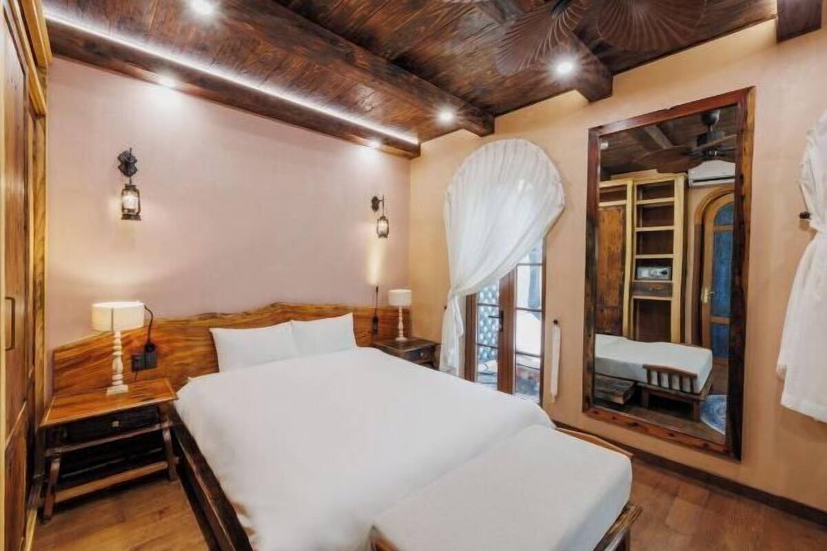 A bedroom with a large bed, wooden headboard, two bedside lamps, a canopy bed, a mirror, and a view of a balcony.