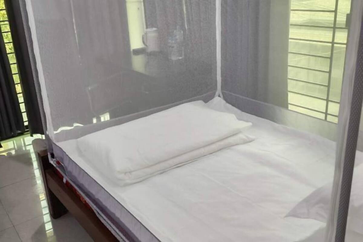 Ein Einzelbett mit weißer Matratze und Kissen, ein Moskitonetz und ein Fenster mit Jalousien.
