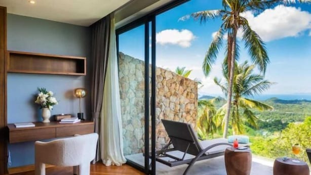 A modern room with a large window, a stone wall, a wooden desk, a white chair, and a lounge chair.