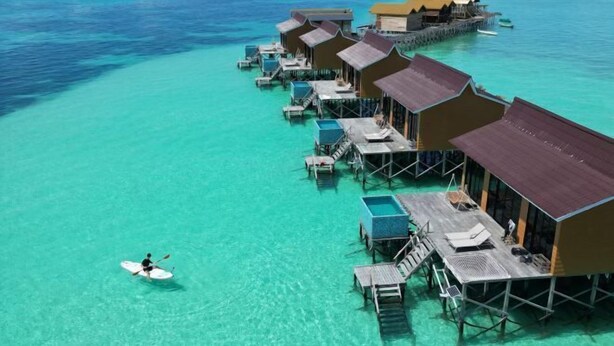 Overwater bungalows with clear turquoise water, a person paddling a paddleboard, and a wooden deck with a table and chairs. - Moana Water Village Semporna (Semporna)