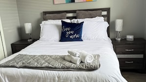 Un letto ben rifatto con lenzuola bianche, un cuscino blu con la scritta 'love you more' e una coperta grigia. Al di sopra del letto, un quadro astratto dai colori vivaci, e un comodino con una lampada e una sveglia digitale.
