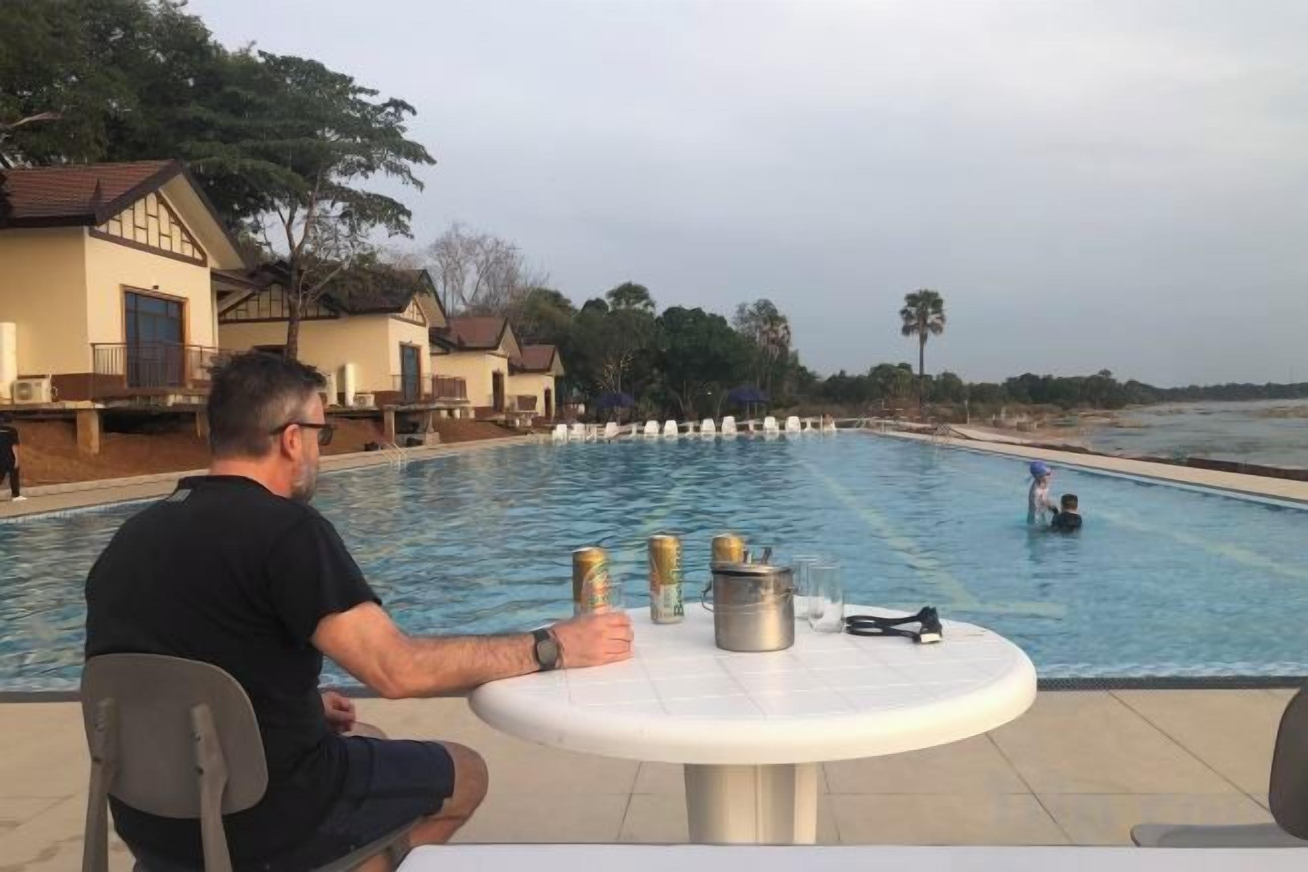 A man sitting by a pool with a table, overlooking a large swimming pool and a beach.