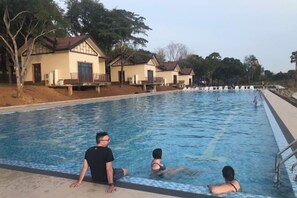 Uma piscina com uma pessoa sentada na borda, outra pessoa na água e uma terceira pessoa ao fundo.