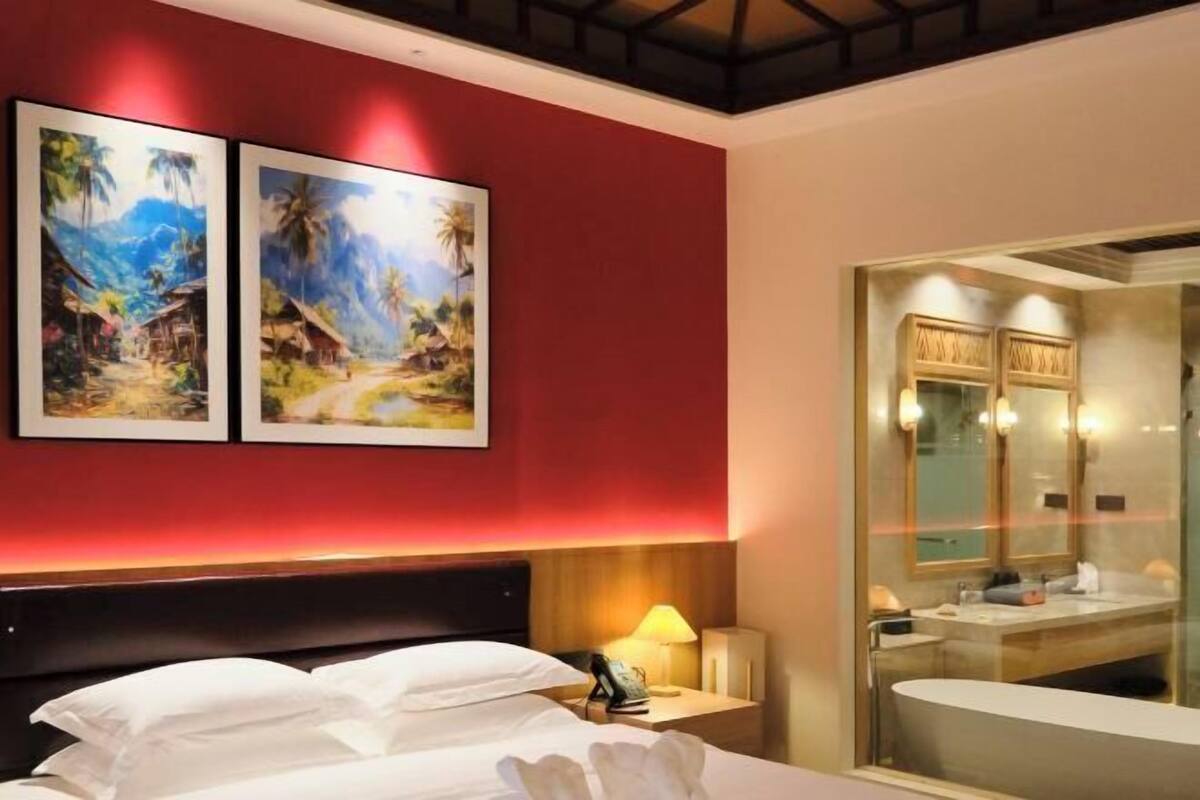 A hotel room with a bed, a nightstand, a lamp, and a large window with a view of the bathroom.
