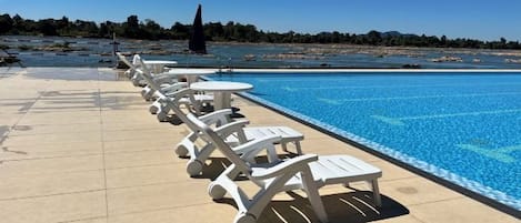 A swimming pool with lounge chairs and tables, a clear sky, and a distant view of the beach.