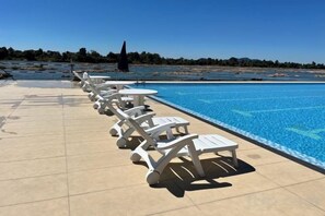 Ein Swimmingpool mit Liegestühlen und Tischen, ein klarer Himmel und ein weiter Blick zum Strand.
