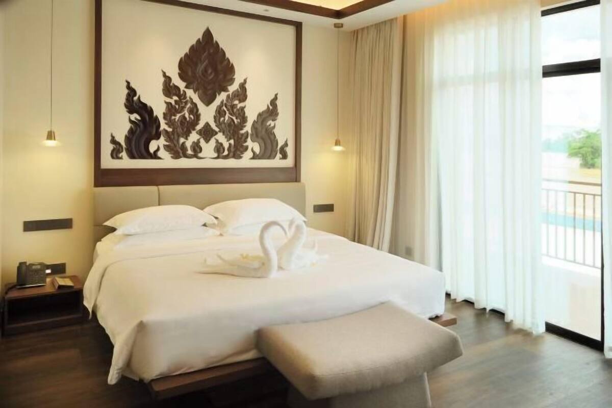A hotel room with a large bed, a decorative wall piece, a bedside table, and a view of the outdoors through a window with curtains.