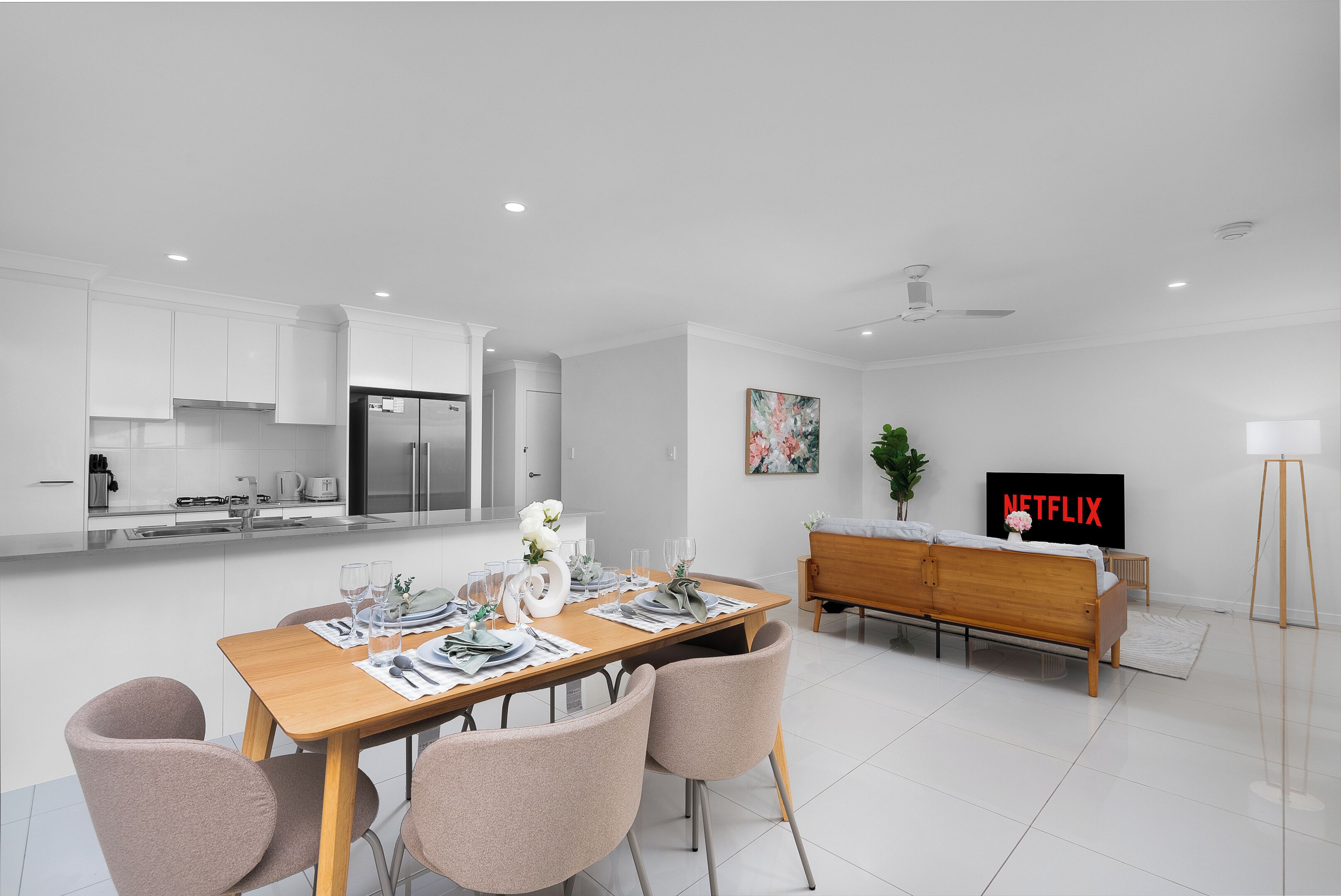 Una cocina moderna con armarios blancos, un comedor con mesa de madera dispuesta para cuatro personas, un sofá y un televisor mostrando 'NETFLIX'.