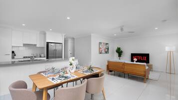 Una cocina moderna con armarios blancos, un comedor con mesa de madera dispuesta para cuatro personas, un sofá y un televisor mostrando 'NETFLIX'.