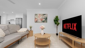 A modern living room with a sofa, a round wooden coffee table, a TV on a wooden stand, and a painting on the wall. - Whispering Pines - Stylish 3BR Duplex close to M1 (Pimpama)