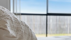 A bed with a white pillow and a view of a wooden fence through a window. - Whispering Pines - Stylish 3BR Duplex close to M1 (Pimpama)