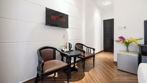 Ein Hotelzimmer mit Holzboden, einem an der Wand montierten Fernseher, zwei Stühlen, einem kleinen Tisch und einer Vase mit Blumen.
