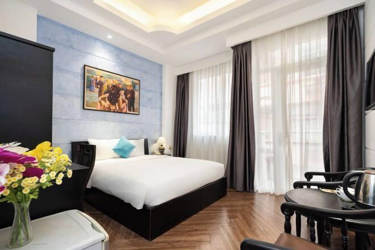 A hotel room with a bed, a desk, a chair, a vase of flowers, a painting on the wall, and a window with curtains.