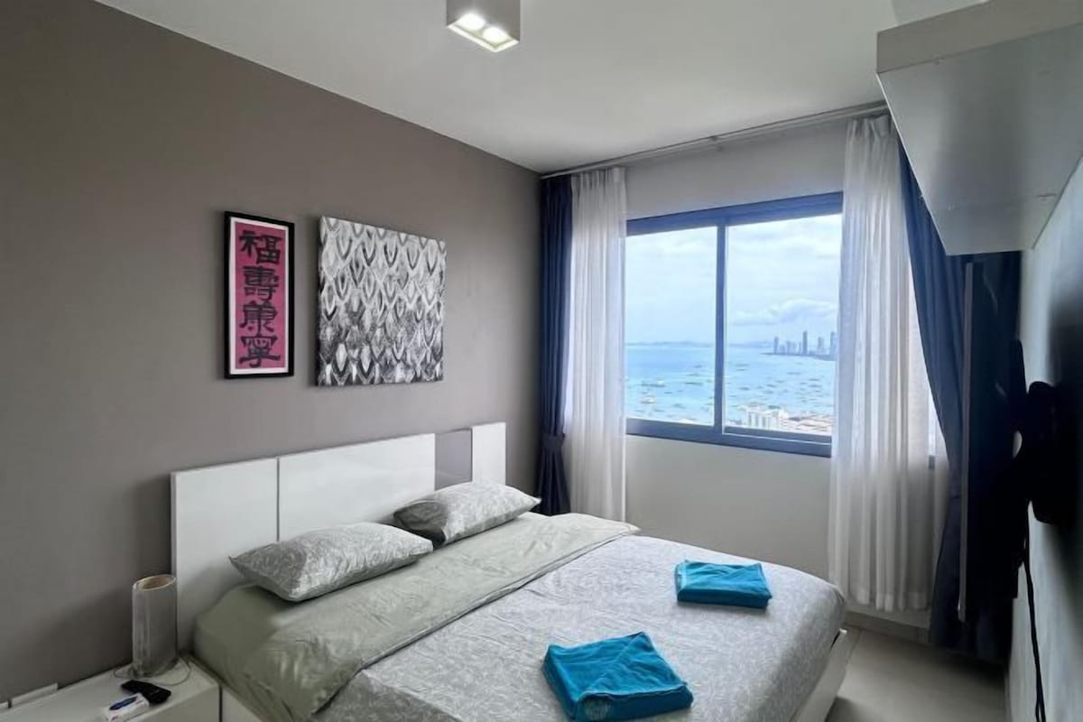 A modern hotel room with a bed, a window with a city view, and artwork on the wall.