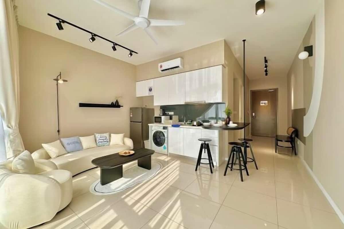 A modern living room with a sofa, a coffee table, a dining area with stools, a kitchen with a microwave, and a ceiling fan.