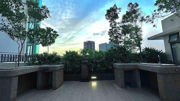 A rooftop terrace with a view of city buildings, a wooden floor, and a central planter with trees.
