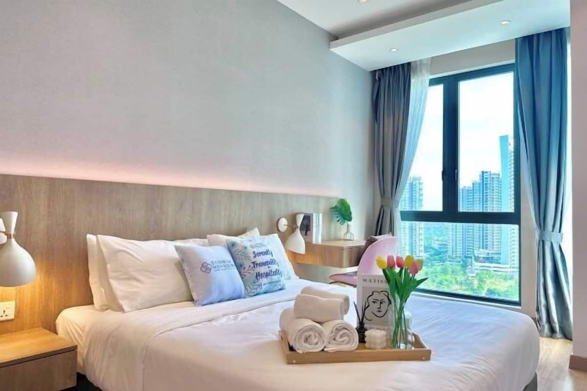 A hotel room with a large bed, a nightstand with a lamp, a tray with towels and a vase of flowers, and a view of buildings through the window.