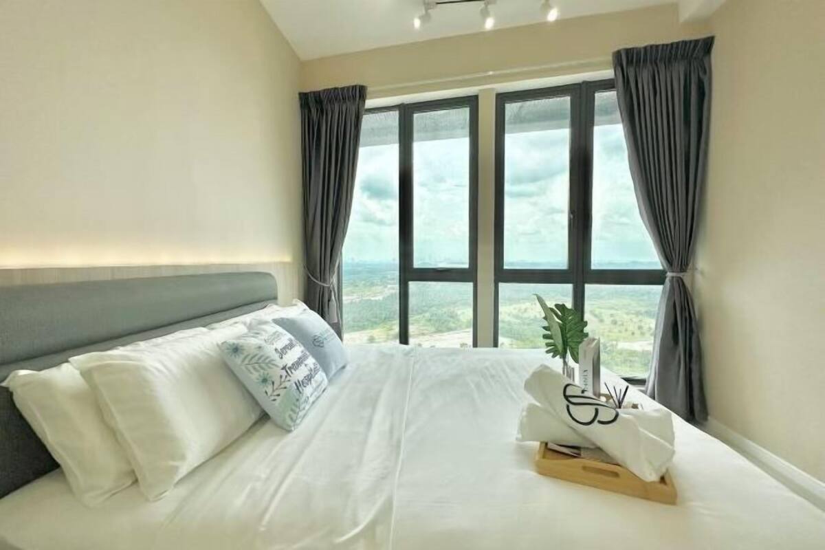 A hotel room with a bed, curtains, a window with a view, and a bedside plant.