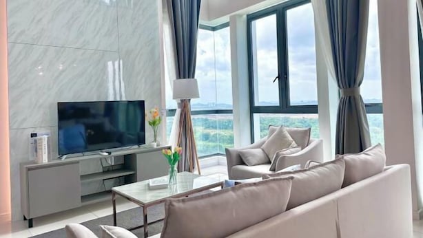 A modern living room with a flat-screen TV, a sofa, a coffee table, and a view of the outdoors through large windows. - Elysia Park Medini by Stayrene near Legoland (Iskandar Puteri)
