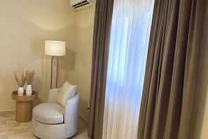 A room with a round white sofa, a wooden side table with a lamp, and a window with curtains. - Victoria (New Cairo City)
