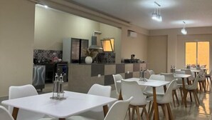 A modern dining area with white tables and chairs, a kitchen in the background, and a large window providing natural light. - Victoria (New Cairo City)