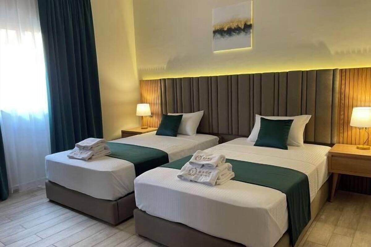 A hotel room with two beds, each with a pillow and a folded towel. There are bedside tables with lamps.