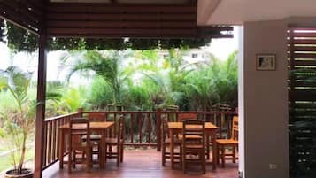 A covered outdoor area with wooden tables and chairs, surrounded by lush greenery.