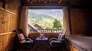 A wooden cabin room with a balcony, a bed, two chairs, a table, and a fan. - Sapa Ta Van Desi House - Mountain View & Hiking Tours (Sapa)