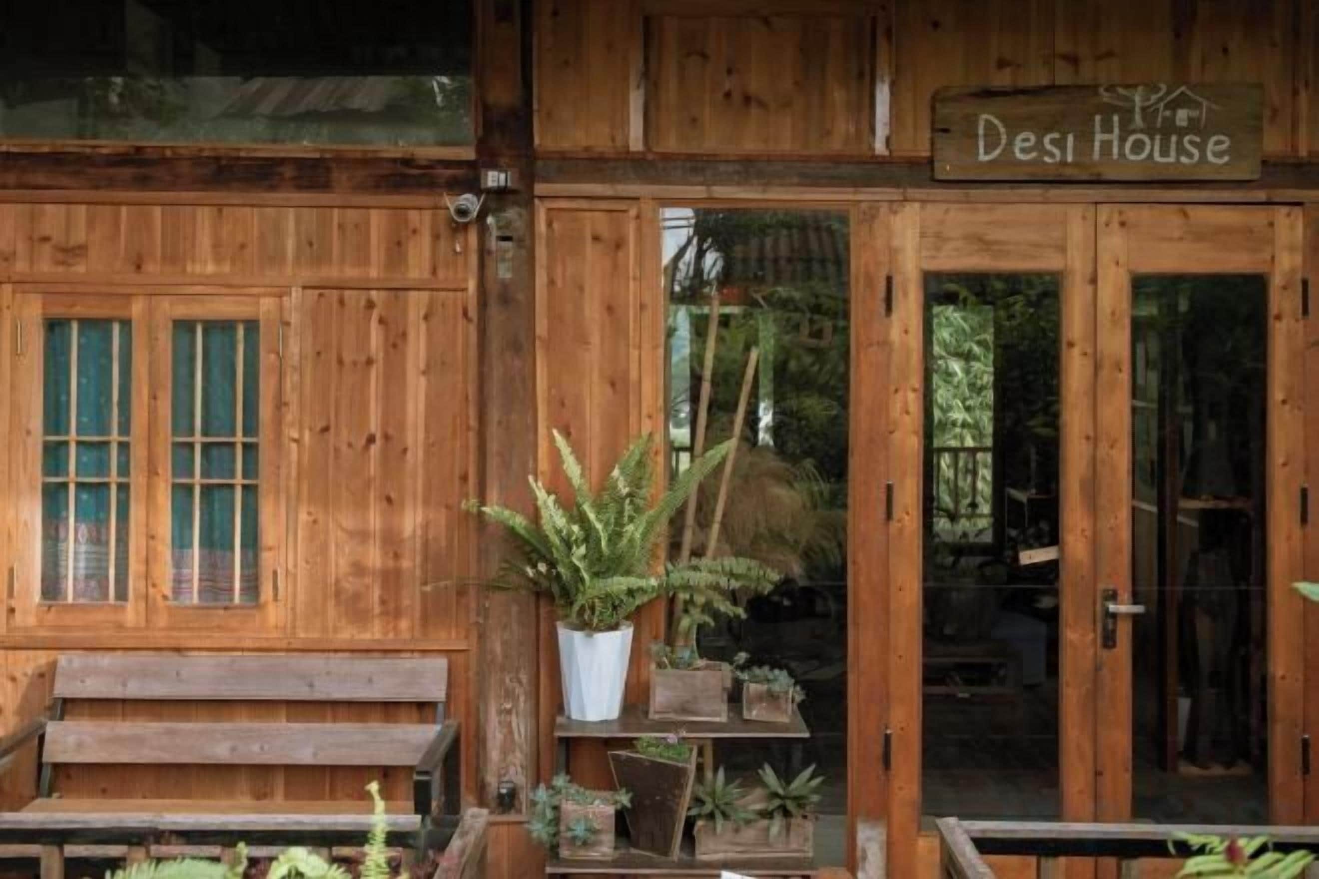A wooden building with a sign reading 'Desi House.' There is a bench outside and several potted plants.