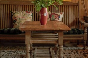 Une table en bois, sur laquelle est posé un vase rempli de plantes vertes, se trouve devant un banc en bois agrémenté de coussins à motifs floraux.