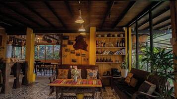 A cozy interior with a wooden ceiling, a patterned floor, a central table with chairs, and a bookshelf filled with books.