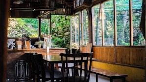 A wooden interior with a stone wall, a table, chairs, and a view of greenery outside. - Sapa Ta Van Desi House - Mountain View & Hiking Tours (Sapa)