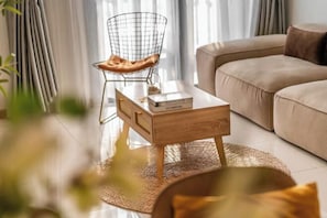 A modern living room with a beige sofa, a wireframe chair with a leather seat, and a wooden coffee table with books.