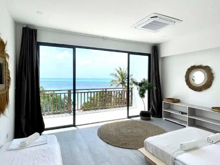 A bedroom with two beds, a large window with a view of the sea, a balcony, and a round mirror on the wall.