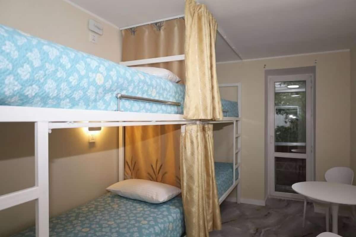 A bunk bed room with a small table and a chair, a window with curtains, and a door leading outside.