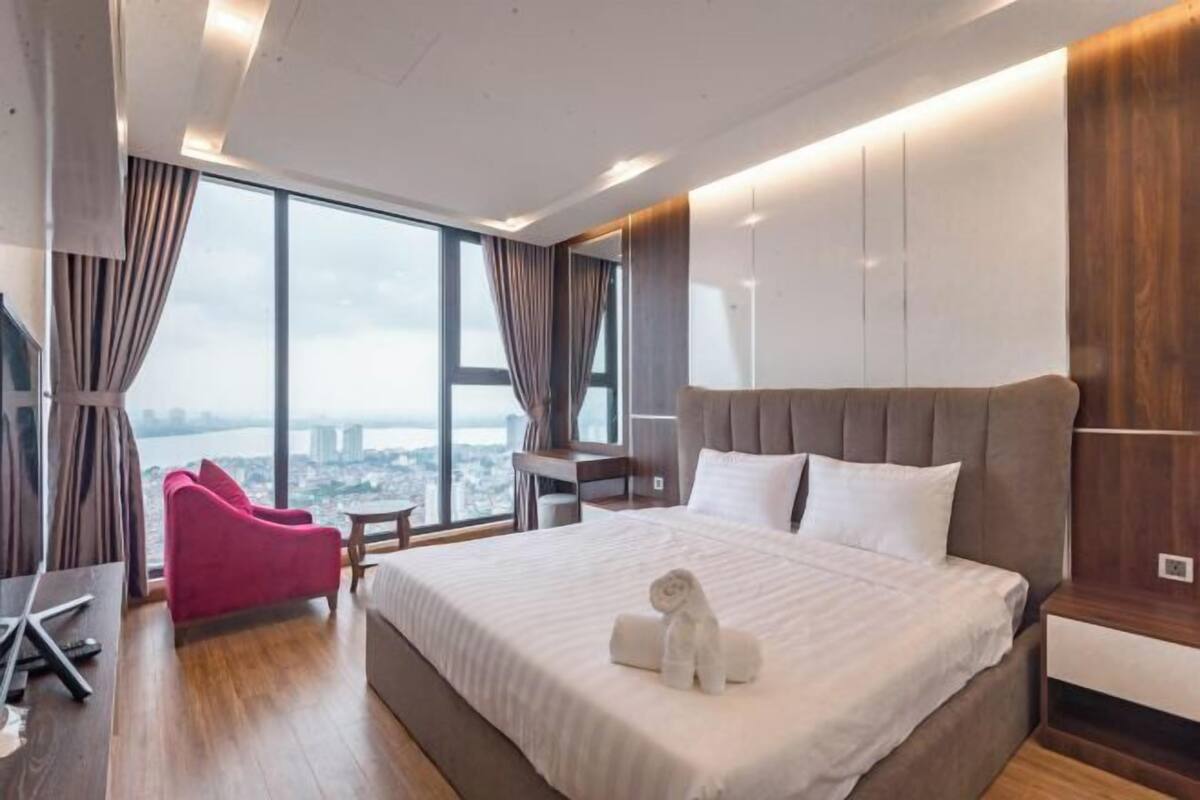 Une chambre d’hôtel moderne avec un grand lit, un fauteuil rouge et une vue sur la ville grâce à de grandes fenêtres.