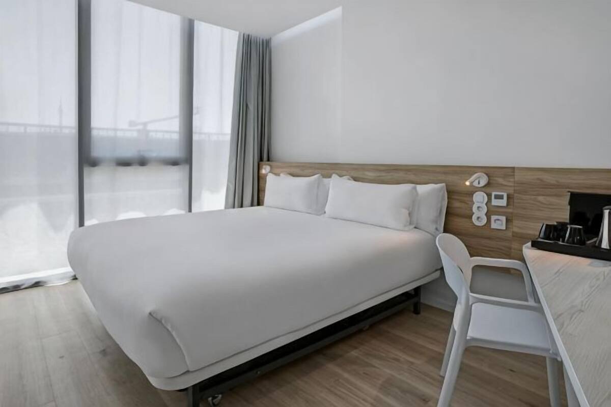 A modern hotel room with a large bed, a wooden headboard, a white chair, and a view of a bridge.