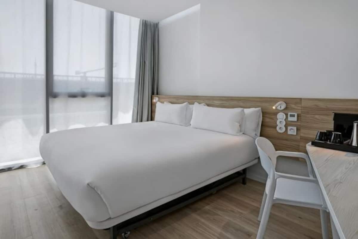 Quarto de hotel moderno com uma cama grande, cabeceira de madeira, uma cadeira branca e vista para uma ponte.