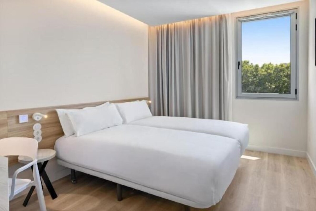 Ein Hotelzimmer mit einem Doppelbett, einem kleinen Schreibtisch mit Stuhl, einem Fenster mit Vorhängen und Blick ins Grüne.