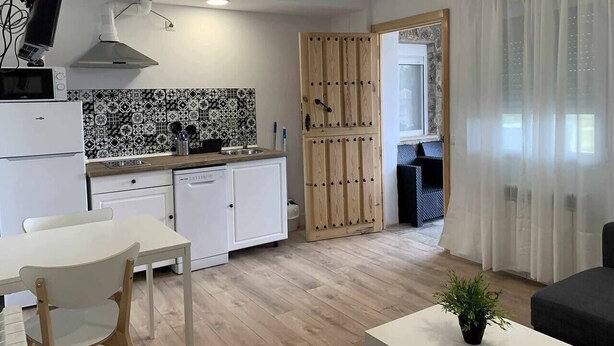 A compact kitchen with a microwave, stove, and sink. There is a dining table with chairs and a small white cabinet. A wooden door leads to another room. - Land. Beautiful apartment. Villa de Gredos (Barajas)