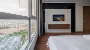 A modern hotel room with a large window, a bed, a mounted TV, and a wooden shelf. - Vinhomes Central Park - PHAN DANG Residences (Ho Chi Minh City)