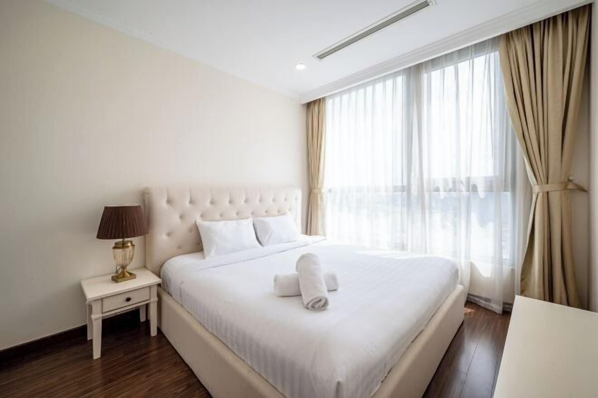 A hotel room with a large bed, a bedside table with a lamp, and a view of the city through large windows.