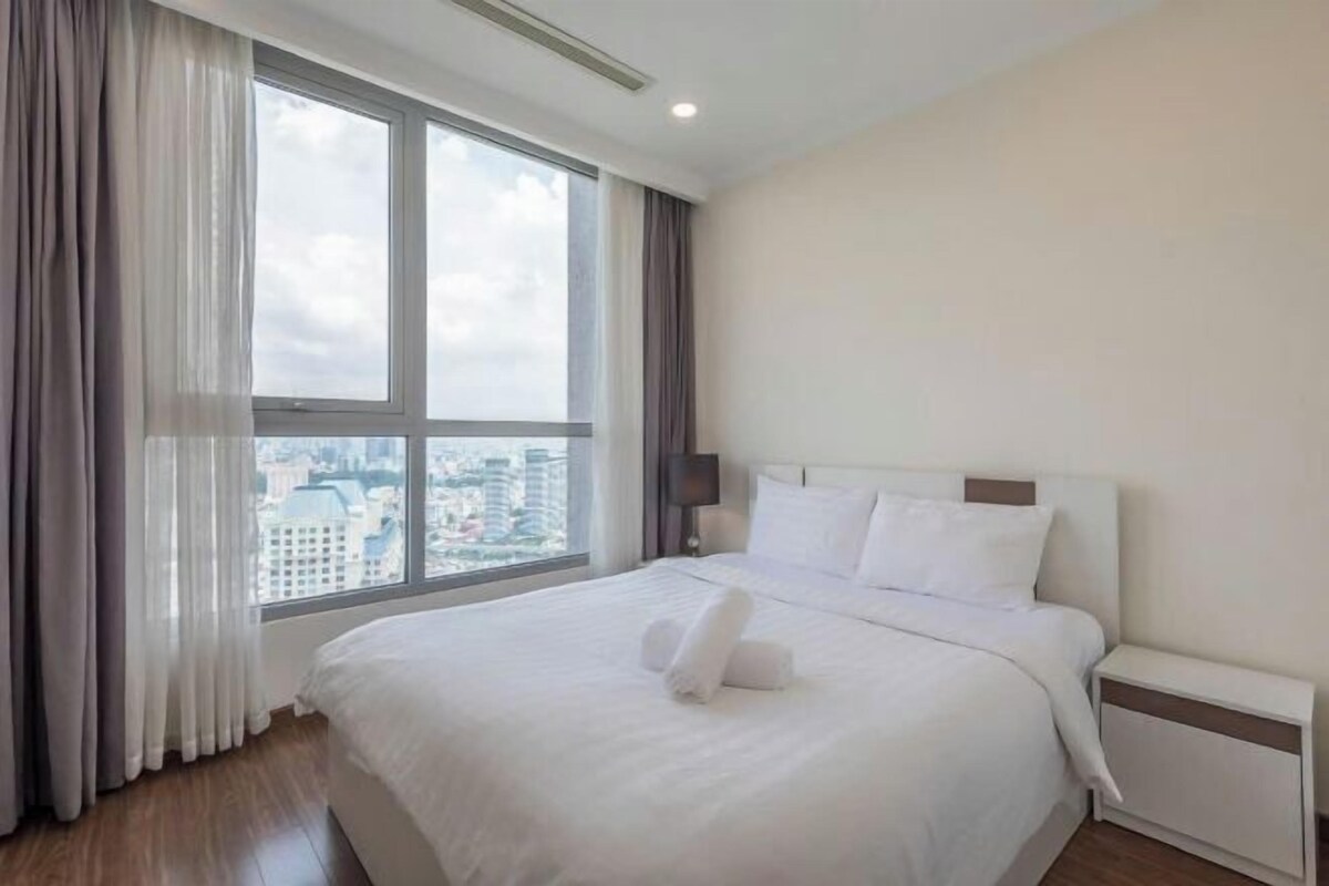 A hotel room with a large bed, a nightstand, a window with curtains, and a city view.
