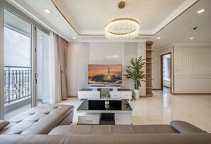 A modern living room with a large window, a chandelier, a flat-screen TV, and a sofa. - Vinhomes Central Park - PHAN DANG Residences (Ho Chi Minh City)