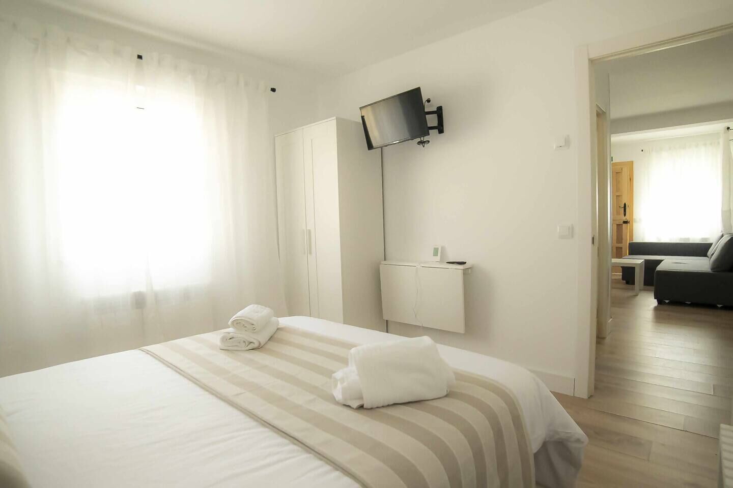 A hotel room with a bed, a TV mounted on the wall, a small bedside table, and a view into the living area.