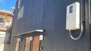 Um edifício com fachada escura, uma porta de madeira e uma pequena caixa elétrica branca na lateral.